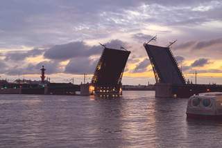 Es ist die Palastbrücke in Sankt-Petersburg zu sehen. Die Brücke öffnet sich gerade, damit ein Schiff die Brücke passieren kann. Im Vordergrund des Bildes sieht man bläuliches Wasser im Hintergrund sind die Gebäude der Stadt zu sehen.