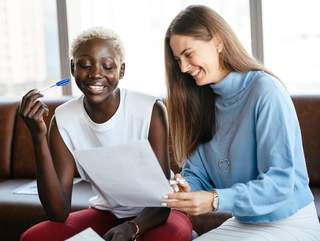 pexels-alexander-suhorucov-6457520 Zwei Frauen sitzen vor einer Fensterfront und schauen gemeinsam auf ein Papier. Die eine Frau deutet mit einem Stift auf eine Stelle im Text, die andere Frau schaut darauf. Sie hat ebenfalls einen Stift in der Hand. Beide Frauen lachen.