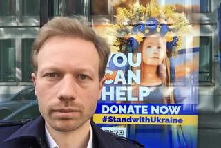 Herr Joschua Ritz steht vor einem Schaufenster. In diesem Schaufenster hängt ein Plakat. Auf diesem Plakat ist ein junges Mädchen zu sehen, dass ein blau-gelbes Kleid und eine blau gelbe Blumenkrone trägt. Auf diesem Plakat steht geschrieben: Du kannst jetzt Spenden. Sei solidarisch mit der Ukraine.